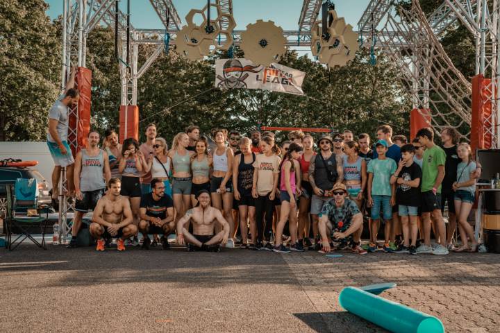 A large group of Ninja Warrior athletes is gathered after a successful 3D Sportanlagen workshop for obstacle parcours in front of the event frame. Above them are various Ninja obstacles hanging, which are mounted to the construction.